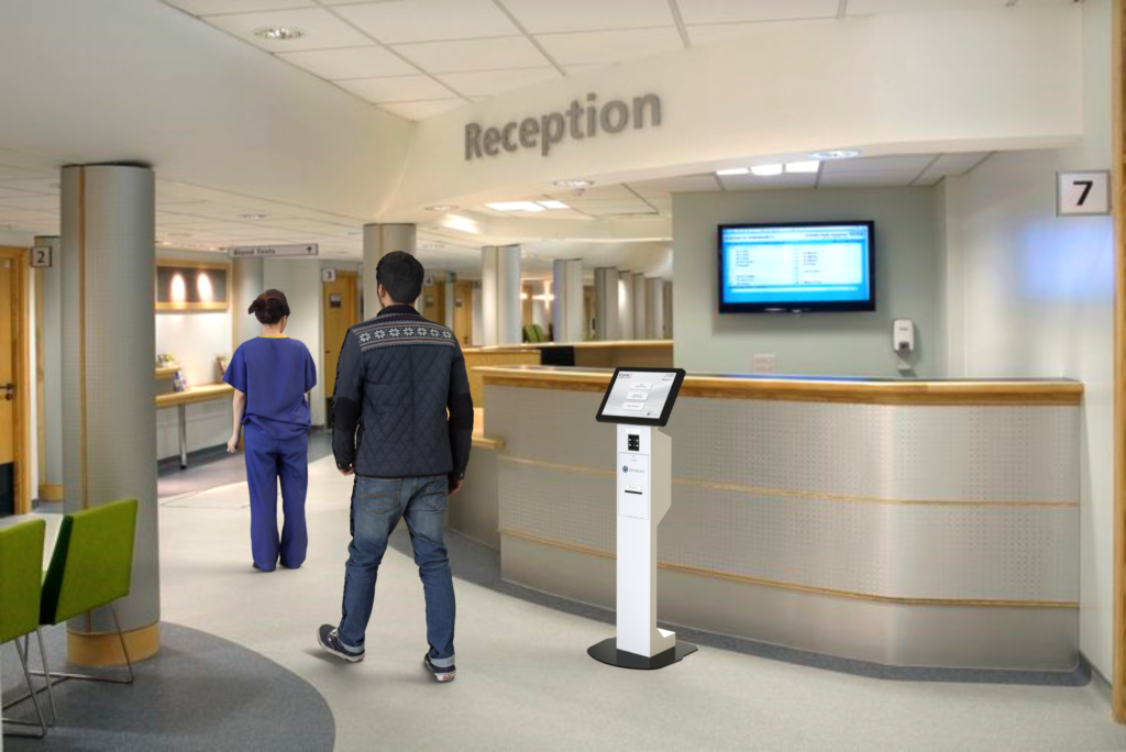 A self-service check-in terminal standing in front of a staffed reception desk in a healthcare facility