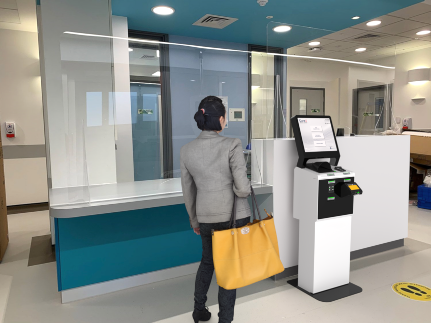A visitor with a shopping bag using a self-service registration terminal beside a service counter